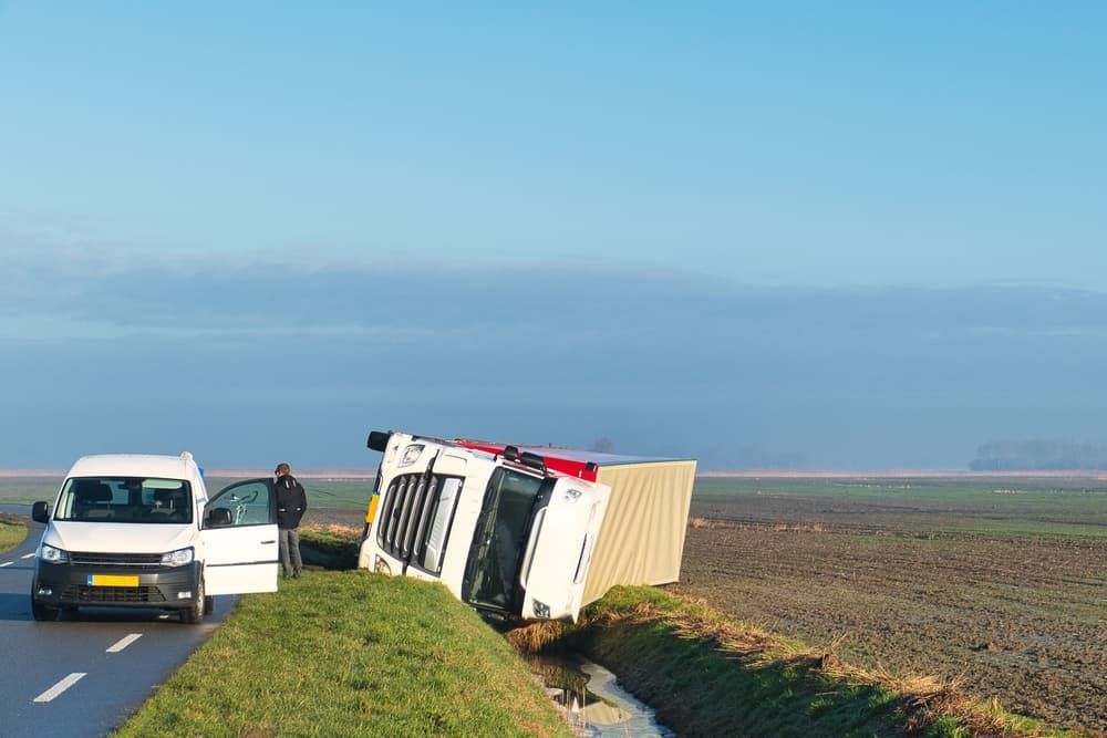 Overturned truck lies in rural ditch; driver stands nearby, awaiting help in isolated farm area.