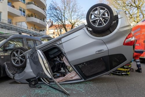 Silver car flipped upside down on a city street after a rollover crash with shattered glass and emergency crew nearby.