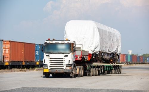 Large flatbed truck transporting a covered oversized load near shipping containers in an industrial area.