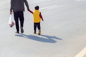 An adult and child holding hands cross a wide street, carrying a bag of red apples.