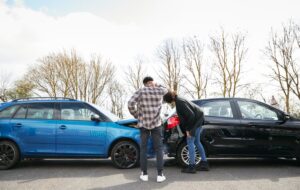 Two people inspect damage after a rear-end collision between a blue car and a black car.