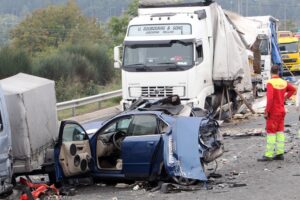 Wreckage from a multi-vehicle truck crash shows crushed cars and emergency responders assessing the scene.