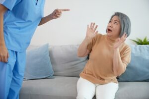 Elderly woman sitting on a couch raising her hands in fear as a healthcare worker stands over her pointing a finger during a confrontation.
