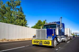 Blue semi truck carrying an oversize load with a bright yellow warning sign drives on a multi-lane highway under a clear blue sky.
