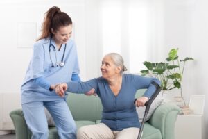 A smiling nurse assists an elderly woman with gray hair as she stands up from a green sofa using a crutch, in a bright living room with plants and natural light.
